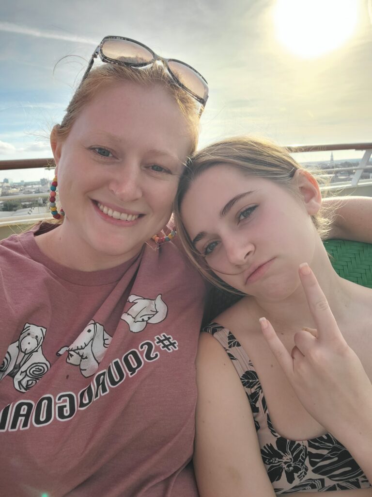 mother and daughter sit on cruise ship deck. mom has arm around girl. girl is making hand gestures and puckering lips, mom is smiling. sun is setting in the background.