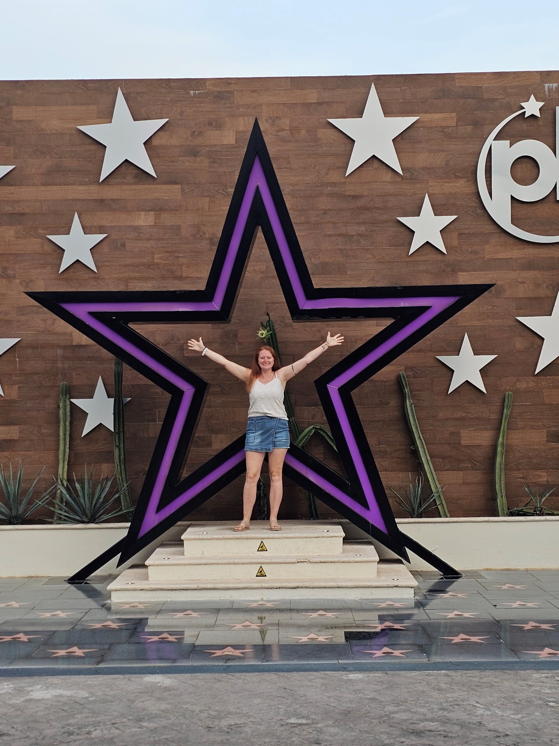 A woman stands on a small platform with arms outstretched in the air. She is smiling. Behind her is a large purple star which is part of the planet hollywood cancun logo
