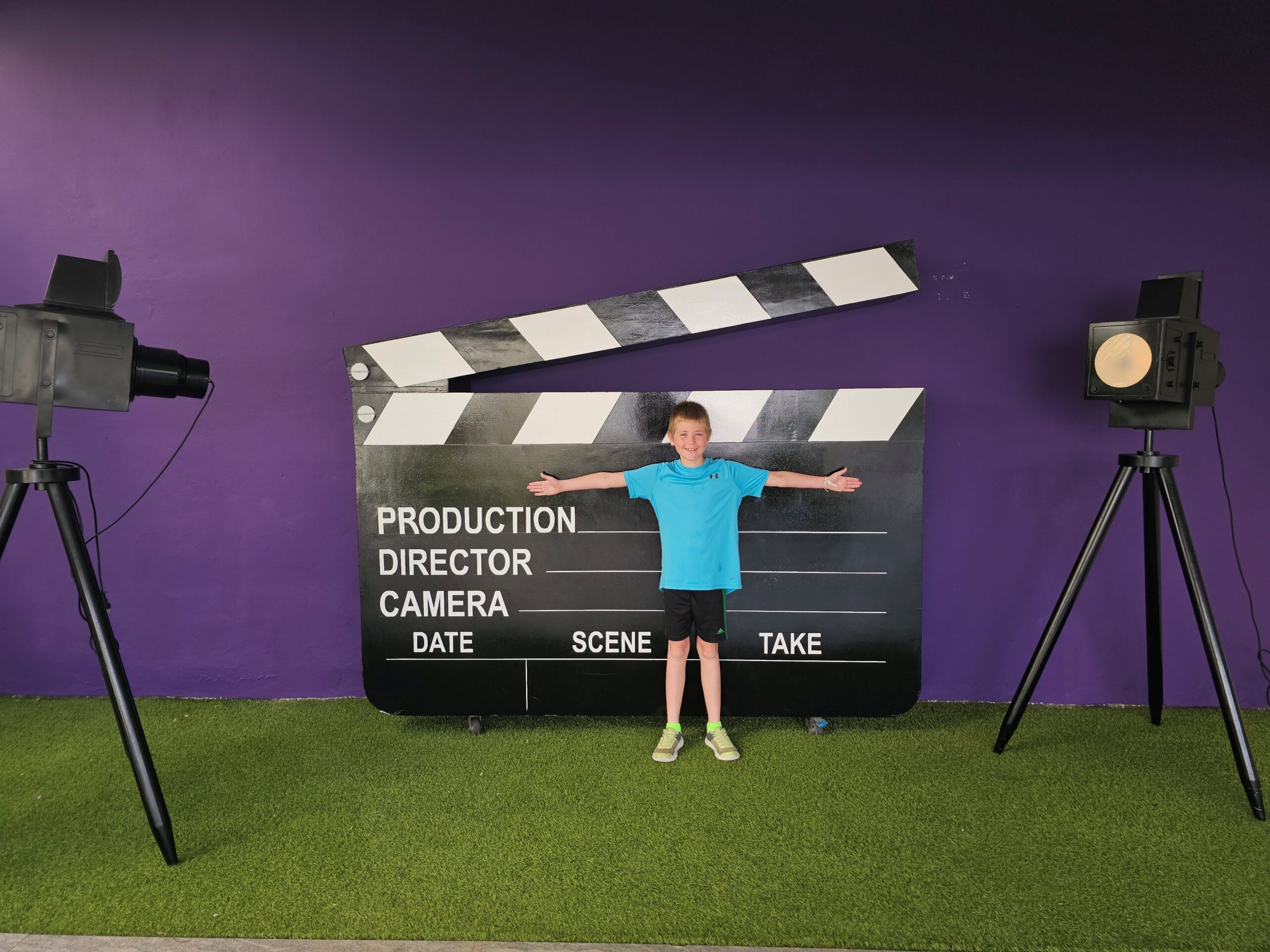 A young boy stand in front of a large prop hollywood director's sign with prop lighting equipment on the sides. He is smiling, wearing a blue shirt and black shorts, and has his arms outstretched across the director's sign. 