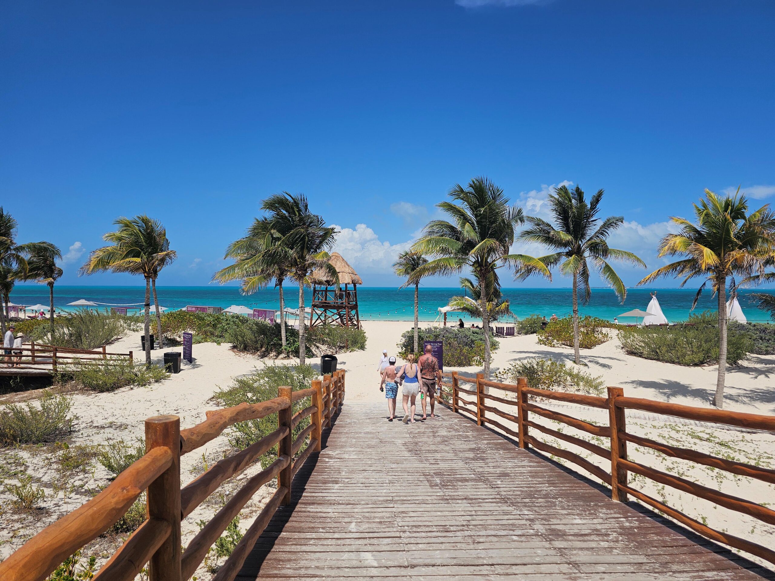 bue ocean water inthe background, in the foreground is a bridge over a sandy beach. A family of a man and some kids walk across the bridge toward the ocean,