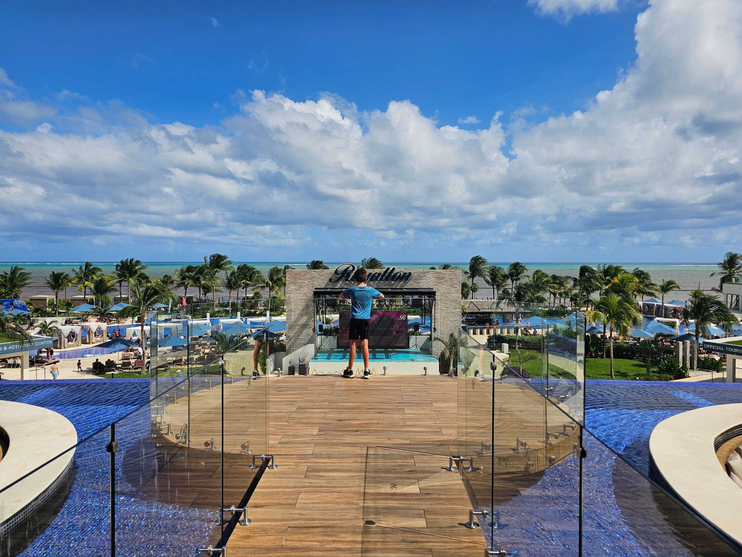a young boy stands on a wooden bridge between two pools overlooking the resort and the ocean beyond.