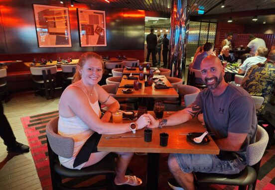 husband and wife sitting at a restaurant holding hands on a cruise ship virgin voyages scarlet lady razzle dazzle lucky lotus