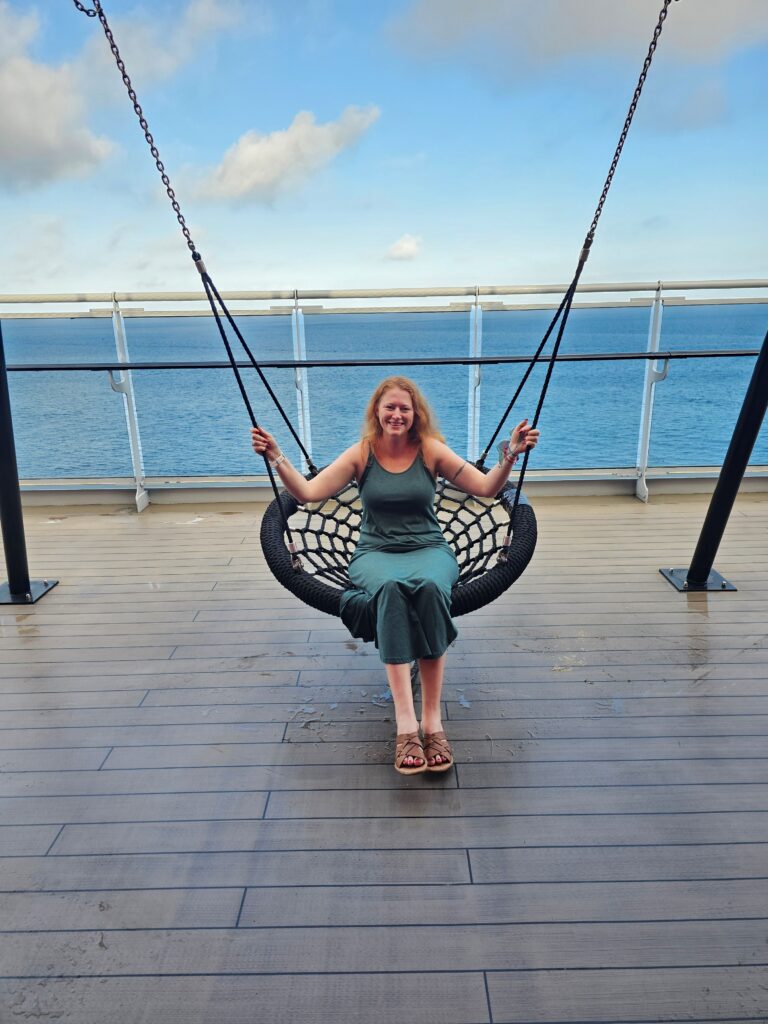 a woman is sitting on a circle swing on the deck of virgin voyages scarlet lady cruise ship with ocean in background