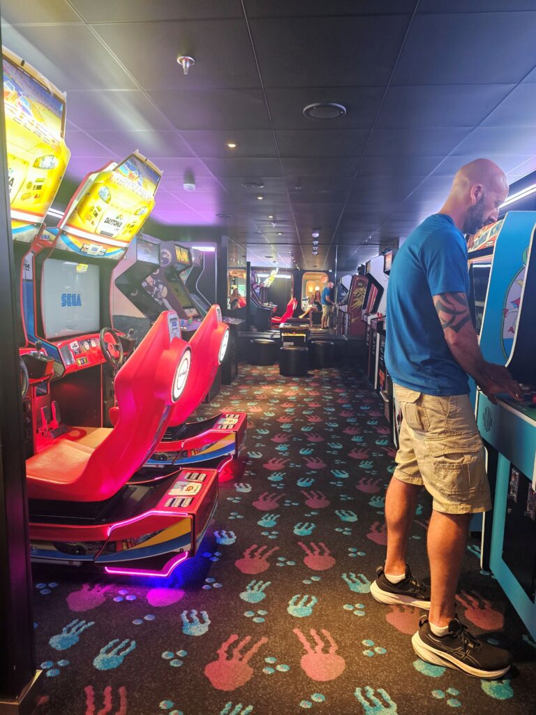 a man playing arcade games in a room full of classic arcade game systems. This is located onbord virgin voyages scarlet lady.