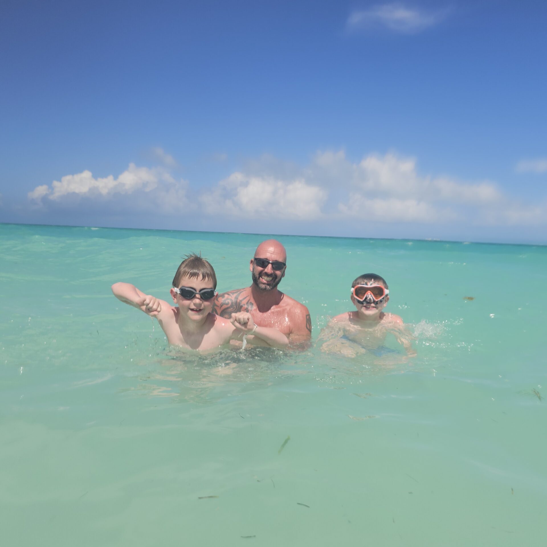 A man and two boys play together in the ocean, the water is somewhat clear and it is a light turquois color. It is a sunny day with only a few clouds in the distance. 
