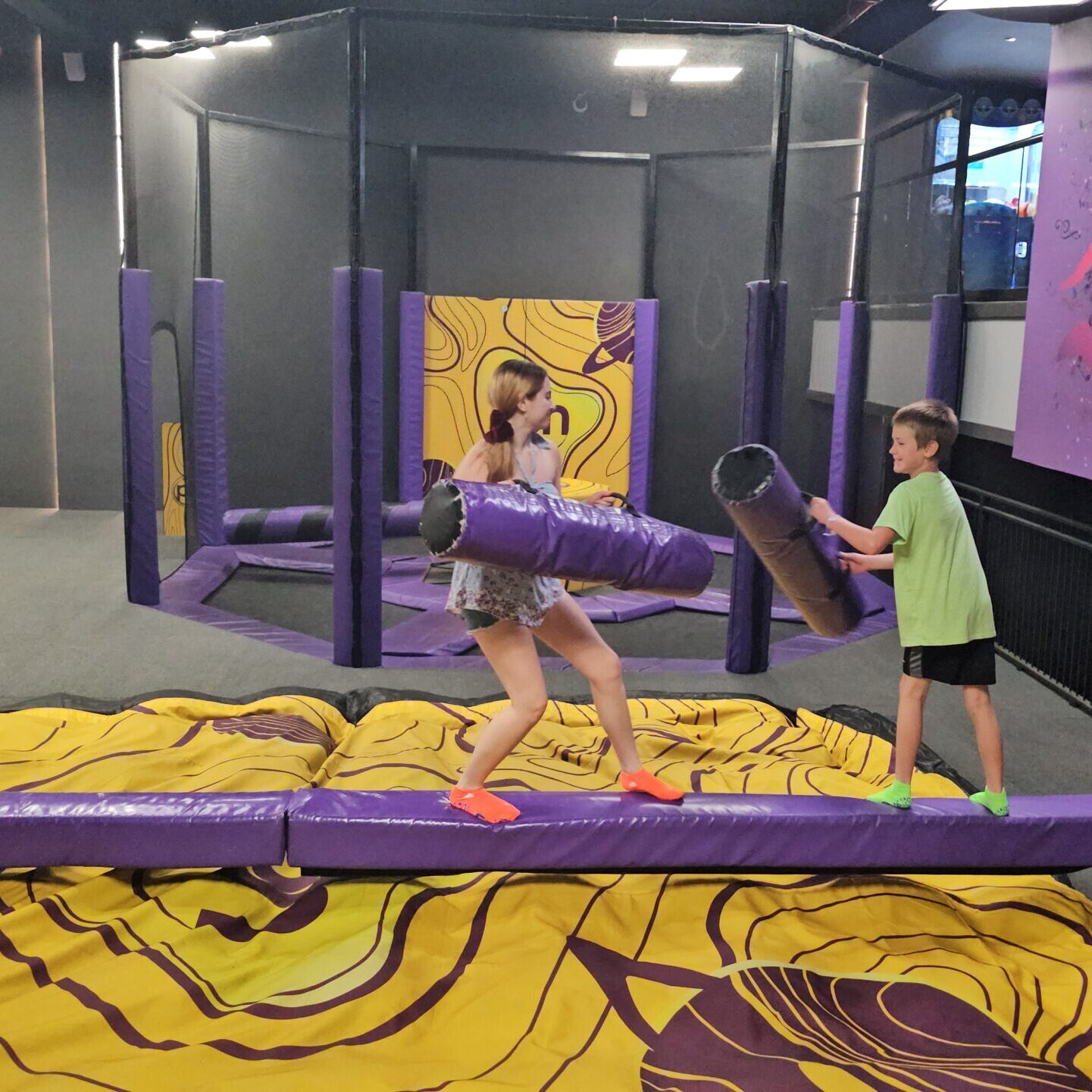 A young boy and teenage girl are staning on a balancing beam at a trampoline park at planet hollywood cancun. They each have foam rolls that they are using to try to knock each other off of the beam.