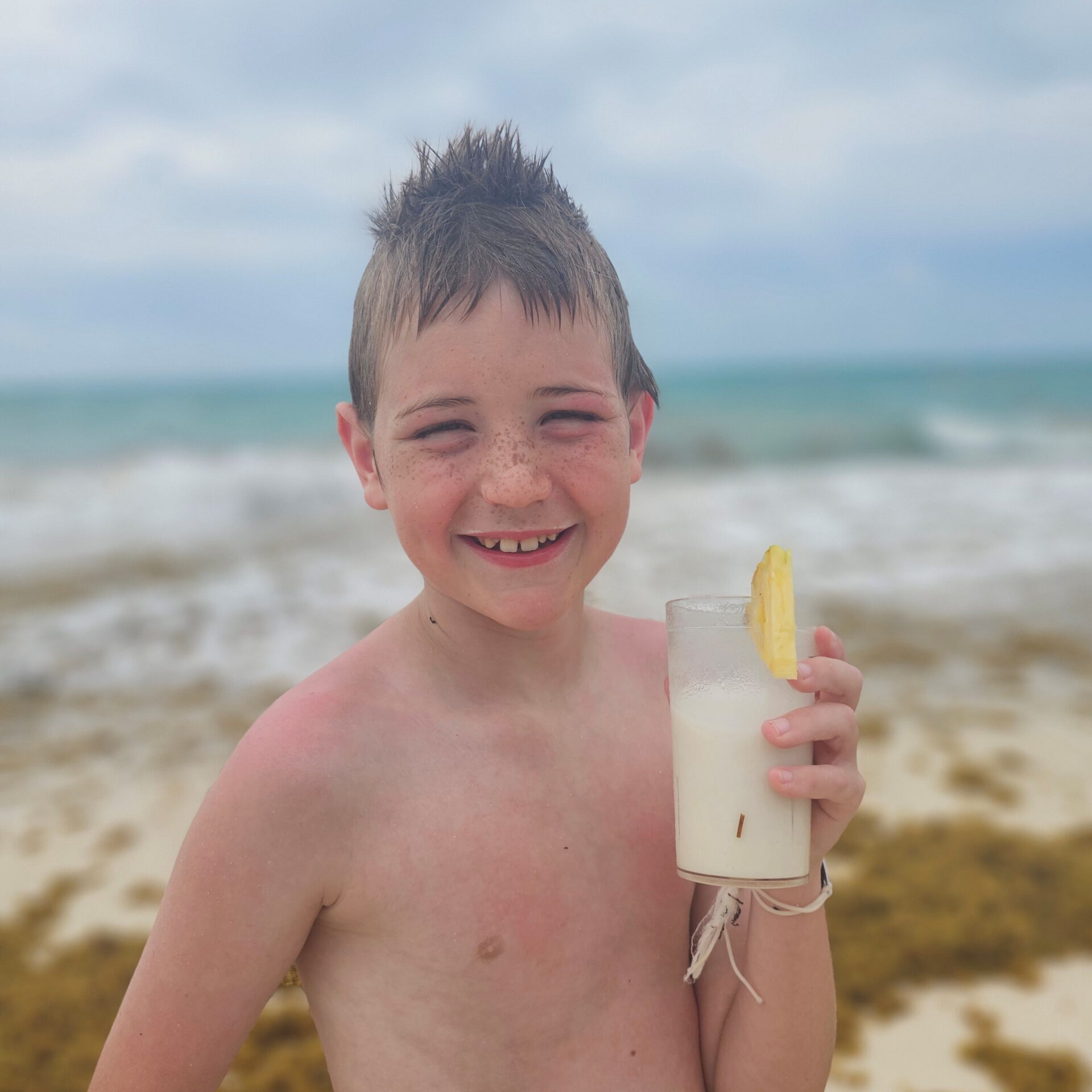 A boy is standing on the beach holding a cup filled with a frozen pina colada and smiling. In the background is the ocean, sand and sargassum seaweed.