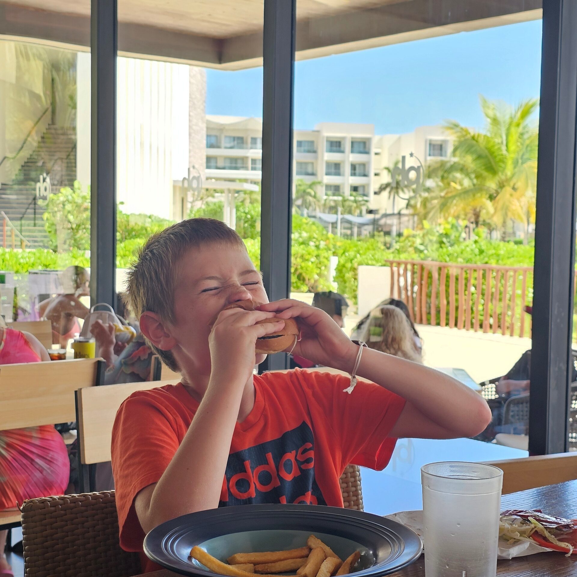 A boy in a bright orange shirt is taking a big bite out of a cheeseburger at the Guy's Burger Joint at Planet Hollywood Cancun