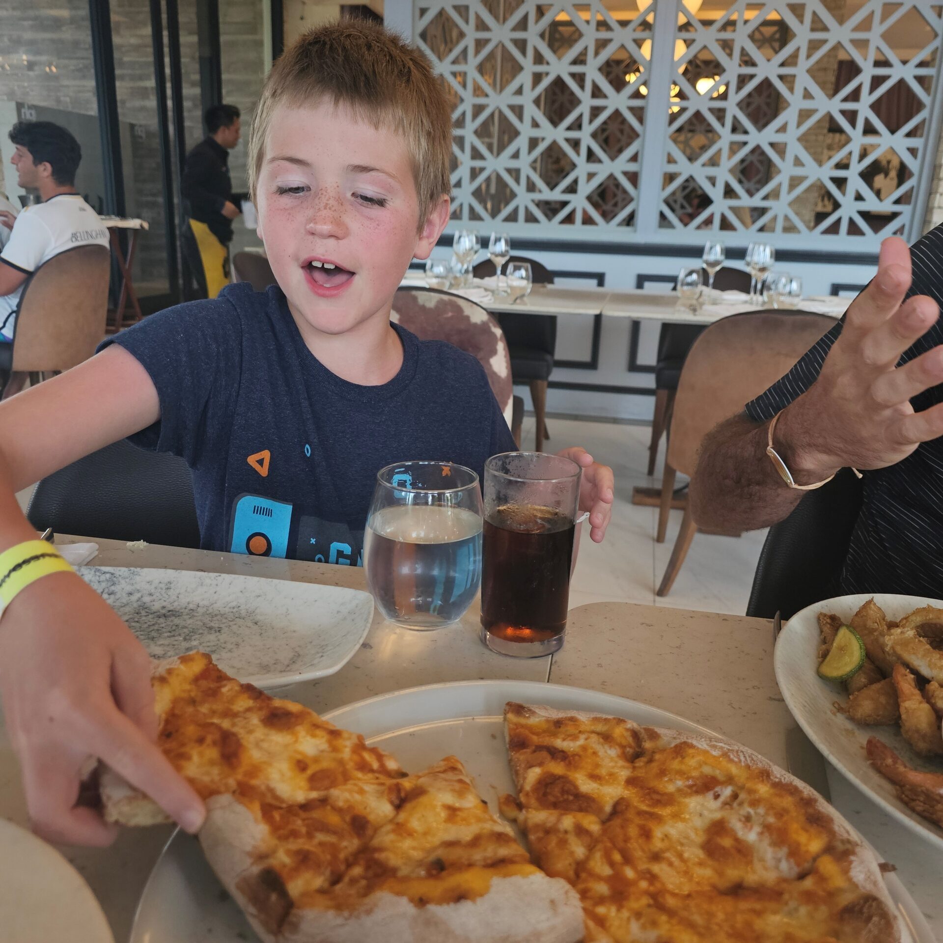 A blonde haired boy is grabbing a piece of freshly baked cheese pizza from a tray on the table in front of him.