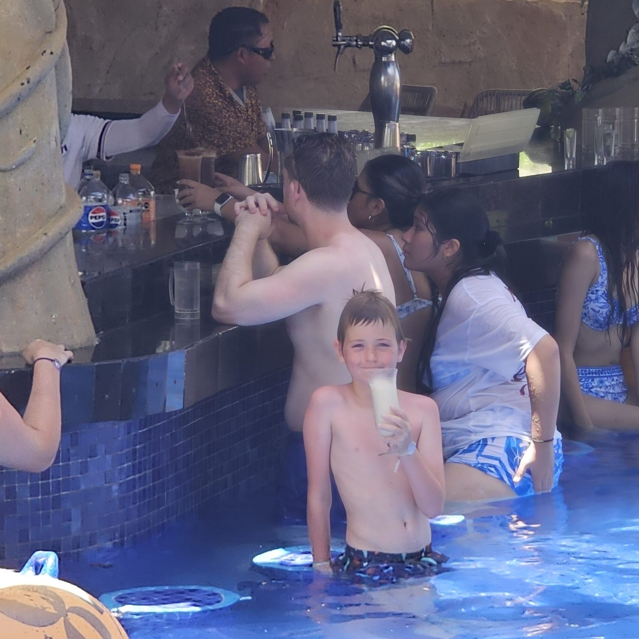A little boy is at a swim up bar in cancun in a lazy river drinking a pina colada and smiling while adults in the background order drinks and swim.
