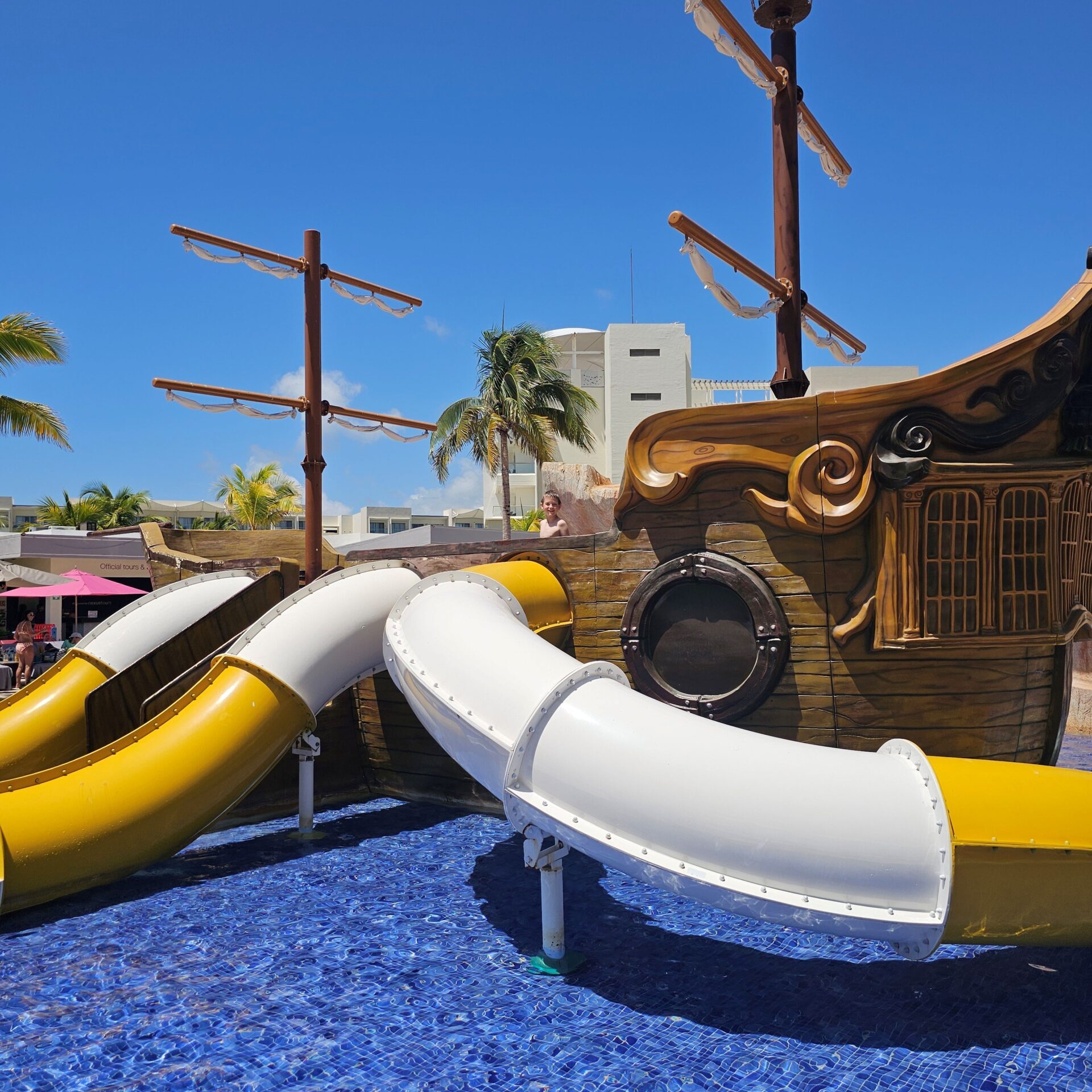 A boy standing on a play pirate ship at a splash park pad at lanet hollywood cancun. The pirate ship has slides that are coming off of the side of it. 