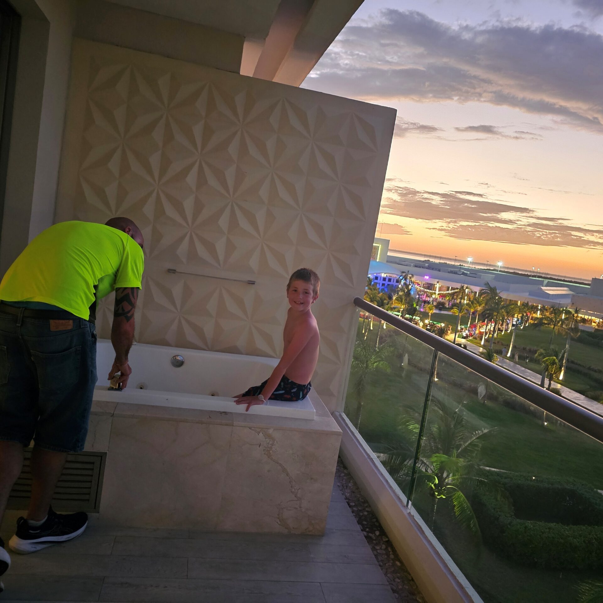 A man is turning on the water to a tub that is on a patio overlooking a resort garden and entertainment area. There is a little boy sitting on the edge of the tub waiting to get in. It is sunset so the sky is darker and lights are on. 